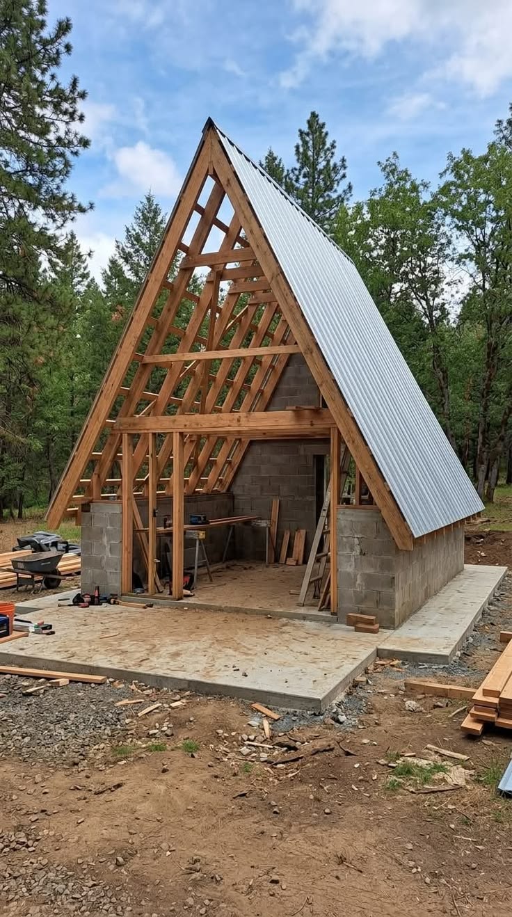 A-frame house under construction showing timber rafters, metal roofing, and concrete foundation in a wooded setting